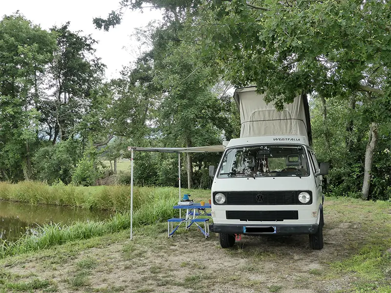 petite-img-tao-3 Image de "Tao" en bivouac en pleine nature au bord d'un plan d'eau en Rhône Alpes. La table et les chaises sont sorties ainsi que le store latérale. Ce van est en location chez Ryôko - spécialiste de la location de vans pour slow travel dans la Loire.